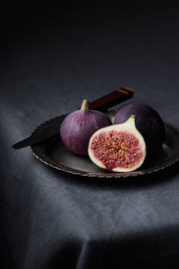 Koyu arkaplanda taze incir, bir parça taze, olgun, lezzetli ve tatlı bir tatlı. Koyu bir masa örtüsü üzerinde mutfak bıçağı, dikey üst görünüm minimalist afiş ve fotokopi alanı.