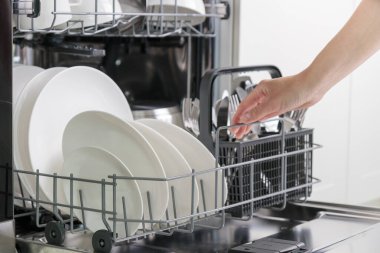 Kadın eli bulaşık makinesini sofra takımıyla dolduruyor. Mutfak aletleri, yaşam tarzı manzarası. Kadın bulaşık makinesinden çatal bıçak sepetini alıyor. Mutfaktaki modern teknoloji kavramı