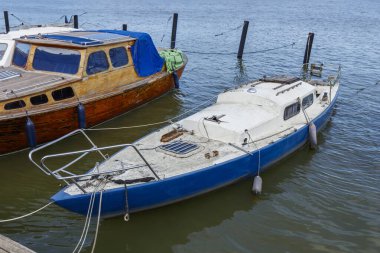 Denizde terk edilmiş, eski, kirli, bakımsız bir yelkenlinin kapısına yakın açık mavi ve beyaz yelkenli, kırık direkli.