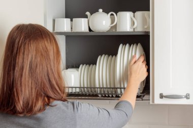 A young woman takes a plate from a dish-drying rack neatly integrated into a wall cabinet in a modern kitchen. The rack holds white ceramic kitchenware, plates and mugs, all arranged orderly. Well-organized aesthetic of simplicity and efficiency clipart