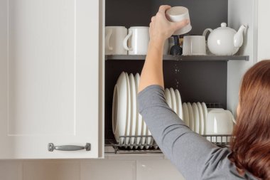 A young woman puts a mug in a dish-drying rack neatly integrated into a wall cabinet in a modern kitchen. The rack holds white ceramic kitchenware, plates and mugs, all arranged orderly. Well-organized aesthetic of simplicity and efficiency clipart