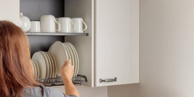 Genç bir kadın, modern bir mutfağın duvar dolabına düzgünce yerleştirilmiş tabak kurutma rafına tabak koyuyor. Askıda beyaz seramik mutfak gereçleri, tabaklar ve kupalar var. Hepsi düzenli olarak düzenlenmiş. Basitlik ve verimliliğin iyi organize edilmiş estetiği
