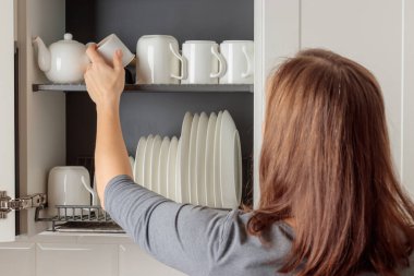 Genç bir kadın, modern bir mutfaktaki duvar dolabına düzgünce yerleştirilmiş bulaşık makinesinin rafına bir kupa koyar. Askıda beyaz seramik mutfak gereçleri, tabaklar ve kupalar var. Hepsi düzenli olarak düzenlenmiş. Basitlik ve verimliliğin iyi organize edilmiş estetiği
