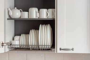 A sleek metal dish-drying rack is neatly integrated into a wall cabinet in a modern kitchen. The rack holds white ceramic kitchenware, plates and mugs, all arranged orderly. Well-organized aesthetic of simplicity and efficiency clipart