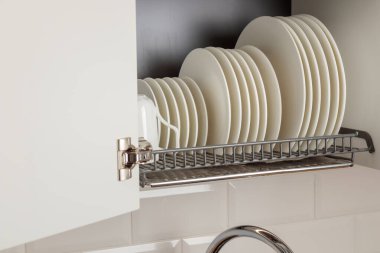 Parlak metal bir bulaşık kurutma askısı modern bir mutfaktaki duvar dolabına düzgünce yerleştirilir. Askıda beyaz seramik mutfak gereçleri, tabaklar ve kupalar var. Hepsi düzenli olarak düzenlenmiş. Basitlik ve verimliliğin iyi organize edilmiş estetiği