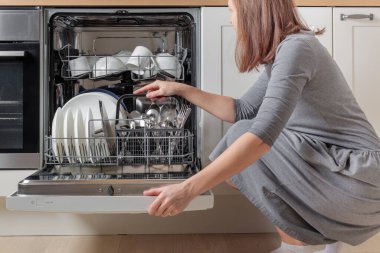 Kadının elleri boşalıyor, bulaşık makinesini boşaltıyor, çatal, kaşık, bıçak ve çatal dolu çatal bıçak sepetini çıkarıyor. Modern mutfak aletleri olan ev işleri. Beyaz tabakları ve paslanmaz çelik çatal bıçakları olan açık bir bulaşık makinesi.