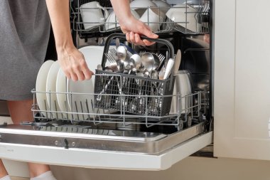 Kadının elleri boşalıyor, bulaşık makinesini boşaltıyor, çatal, kaşık, bıçak ve çatal dolu çatal bıçak sepetini çıkarıyor. Modern mutfak aletleri olan ev işleri. Beyaz tabakları ve paslanmaz çelik çatal bıçakları olan açık bir bulaşık makinesi.