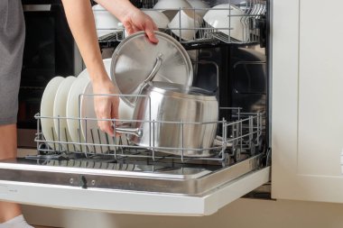 Kadının elleri boşalıyor, bulaşık makinesini boşaltıyor, su ısıtıcısını çıkarıyor. Modern mutfak aletleri olan ev işleri. Beyaz sofra takımı olan açık bir bulaşık makinesi.