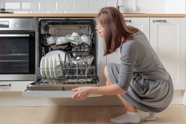 Bir kadın açık bir bulaşık makinesine bakıyor. Temiz, yıkanmış beyaz bulaşıklar ve paslanmaz çelik çatal bıçak takımı beyaz, minimalist bir ev mutfağında. Ev işleri için kullanışlı modern bir araç.
