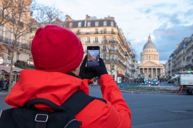 Kırmızı ceketli yalnız bir turist, Paris, Fransa 'daki akıllı telefonuyla büyük neoklasik mimarisini ve ebedi güzelliğini yakalıyor.