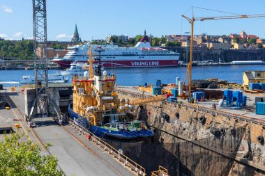 Stockholm, İsveç - 9 Temmuz 2025: Beckholmen adasındaki tarihi bir rıhtımın güneşli geniş manzarası içinde büyük bir gemi ve arka planda açık mavi gökyüzünün altında görünen bir yolcu gemisi
