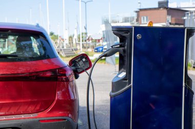 Bir şarj istasyonuna park edilmiş kırmızı elektrikli araba, fişe takılı ve elektriği şarj ediyor, temiz enerji ve sürdürülebilir şehir ulaşımı sergiliyor