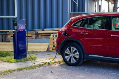 Bir şarj istasyonuna park edilmiş kırmızı elektrikli araba, fişe takılı ve elektriği şarj ediyor, temiz enerji ve sürdürülebilir şehir ulaşımı sergiliyor