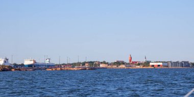 Hanko şehrinin denizden panoramik görüntüsü, tarihi kırmızı su kulesi, Lüteryen Katedrali, ve gemilerle dolu liman, kıyı yaşamı ve mimari simgeleri ön plana çıkarıyor.
