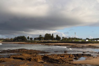 Dramatik bulutlu bir gökyüzü, kayalık bir kıyı şeridi ve kıyıya vuran dalgaların yer aldığı manzaralı bir kıyı manzarası. Uzakta ağaçlar ve park halindeki arabalar görülebiliyor. Güneş batarken sakin ama kasvetli bir atmosfer yaratıyorlar..
