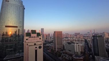 Güneş yüksek gökdelenlerin arkasında batıyor, şehrin üzerine sıcak bir parıltı saçıyor. Manzara modern mimarinin ve kentsel yaşamın canlı atmosferinin bir karışımını sergiliyor..
