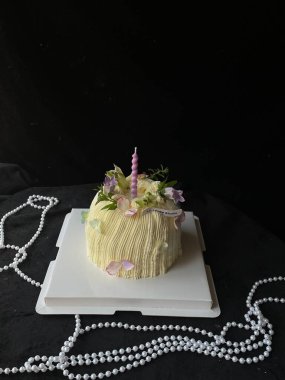 Güzel dekore edilmiş bir pasta, taze çiçekler ve tek bir mum ile süslenmiş beyaz bir kutunun üzerinde oturur. Pastayı inciler sarar, özel bir durum için şenlikli atmosferi güçlendirir..