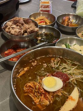 Lezzetli bir Kore soğuk şehriye tabağı büyük bir kasede sunulur, yanında sığır eti, salamura sebze ve deniz yosunu gibi geleneksel tatları ön plana çıkaran bir dizi meze bulunur..