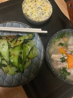 Konforlu bir ev yemeği bir kase içinde kızarmış sebzeler, bir yandan pirinç ve rahatlatıcı bir tavuk çorbası sergiler. Bu yemek sıcaklığı ve aile birlikteliğini yansıtıyor..