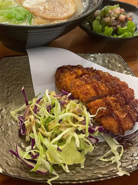 Lezzetli bir tabakta taze marul ve lahana yatağında dinlenen çıtır ekmekli tavuk, yanında da bir kase haşlanmış ramen ve küçük bir meze bulunuyor..