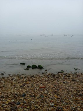 Kum ve kayalardan oluşan bir şerit, sakin bir deniz, uzakta küçük tekneler, bulutlu gökyüzü ve sisli bir gün..