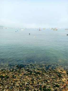 Kayalardan oluşan bir şerit, yeşil renkli berrak bir deniz, sakin, küçük tekneler, bulutlu ve sisli berrak bir gökyüzü. 