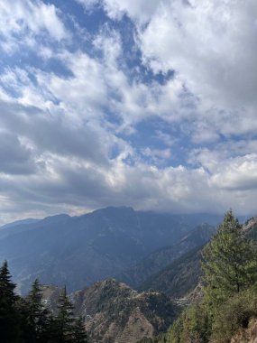 Kısmen bulutlu bir gökyüzünün altındaki dağ sırasının nefes kesici görüntüsü. Uzakta yükselen zirveler, bulutlar başımızın üstünde sürüklenirken yumuşak ışıkla yıkanır. Sahne durgun, uzun yeşil ağaçlar resmi çerçeveliyor..