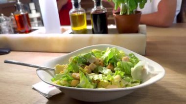 Beyaz kase Sezar salatası masanın üzerinde, çocuklar hareketli pizzacı sahnesinin arka planında oynarken taze tatları eğlenceli, aile dostu atmosferle harmanlıyor. Fesleğen, zeytinyağı
