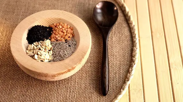 stock image Wooden bowl holding chia seeds, oats, black sesame, lentils paired with wooden spoon on woven mat. Simple, eco-friendly arrangement showcasing sustainable, plant-based nutrition choices Wooden tools