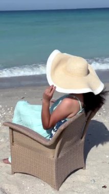 Plaj sandalyesinde uzun siyah saçlı, güneş şapkası takan bir kadın. Deniz kenarında güneşlenmek, tatil ruhunu kucaklamak. Mükemmel seyahat rahatlama sahnesi, gösterişli eğlence ve sahil kenarı rahatlığı.