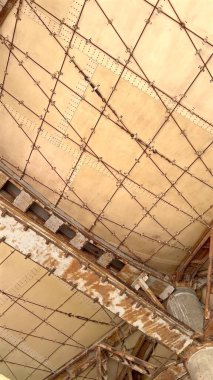 Merkezi kolon, terk edilmiş endüstriyel binada geometrik tavan yapısını oluşturan paslı kirişleri destekliyor. Yıpranmış yüzeyler, çürümüş elementler ve izolasyon uzun vadede terk edilmeyi vurguluyor.