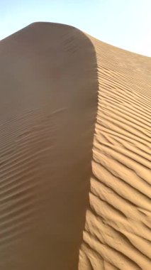 Akşam güneşi, diğer taraf gölgede kalırken yükselen kum tepesinin bir tarafını aydınlatır. Aydınlık ve karanlık arasındaki zıtlık, kumun zarif kıvrımlarını vurgular ve çarpıcı bir çöl manzarası yaratır..