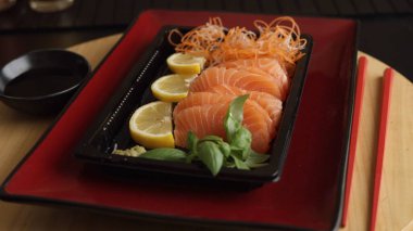 Taze limon ve fesleğenle servis edilen saşimi doğal tatların dengesini vurgulayarak görsel lezzet yaratır. Kırmızı tabak sunumu geliştirirken soya sosu ve özel çubuklar yemek deneyimini tamamlar.