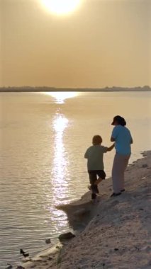 Anne ve oğul suyun kenarında durur altın gün batımının yüzeyde yansıması huzur dolu bir an yaratır batan güneşin ılık parıltısı sessiz bağlantıyı güçlendirir paylaşılan büyülü altın saat