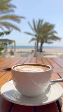 Arka planda bulanık avuç içleri olan plajdan kapuçino. Huzurlu ve rahatlatıcı sahile bakan kahve anları için mükemmel. Doğanın sükunetiyle nefis birayı harmanlıyorlar..