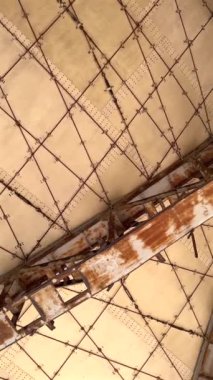 Paslı kirişlerle çöl yapısı, geometrik tavan ve yıpranmış paneller endüstriyel bozulma belirtileri gösteriyor. Dağınık bitkilerle dolu açık kat, uzun süreli terk edilme ve ıssızlığın altını çiziyor.