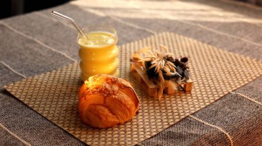 Kruvasan, ekmek tohumu ve örülmüş hasırın üzerine yerleştirilmiş bir bardak sarı meyve suyu sıcak, davetkar bir kahvaltı sahnesi oluşturur. Taze, sanatsal yiyeceğe odaklanmak rahat ve rahat bir sabah yemeği önerir..