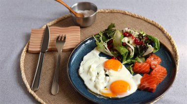 Bright sunny eggs with orange yolks beside green salad, pomegranate seeds, and salmon slices on blue plate, served with silver bowl of salt, emphasizing eco-conscious healthy lifestyle. clipart