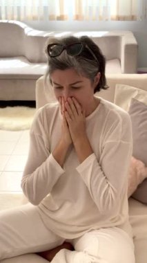 Beyaz elbiseli orta yaşlı bir kadın, bej bej bir zeminde oturuyor, aşağı bakıyor, odaklanmış ruh hali, gri saçlar, doğal ışık huzur ortamını arttırıyor, başındaki gözlükleri takıyor..