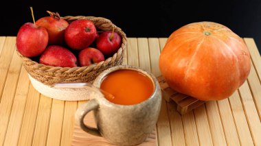 Seramik bardakların yanında kırmızı elmalarla dolu sepet balkabağı, bambu hasırının üzerine yerleştirilmiş balkabağı, güz hasat sahnesi taze ürünler, doğal dokular ve ılık mevsimlik tonlarda sergileniyor..
