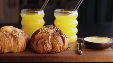 Two croissants placed beside two yellow drinks in glasses with straws resting on wooden board with butter knife. Scene highlights refreshing beverage and fresh pastry pairing for relaxed breakfast.