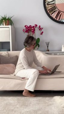 Beyaz kadın bej koltukta oturuyor, laptopta çalışıyor, çiçekler, gri saçlar..