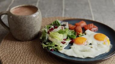 Parlak turuncu yumurtalar, salata, somon ve nar tohumlarıyla birlikte, siyah kahveyle servis edilen, eko-bilinçli yemenin vurgulandığı, renkli sağlıklı kahvaltı sahneleri oluşturur..