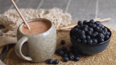 Taze koyu yaban mersiniyle dolu mavi seramik kâse gri fincanın yanında örülmüş bej bir paspasın üzerinde, açık kahverengi smoothie içeriyor, ekolojik bilinçli sağlıklı vegan kahvaltısını vurguluyor..