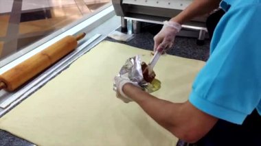 Tarçın dolgusu mavi gömlekli işçiler tarafından düz hamura yayılıyor, fırın üretim ortamında lezzetli tarçın ruloları yaratmak için hassasiyet ve tekniğe odaklanıyor. 
