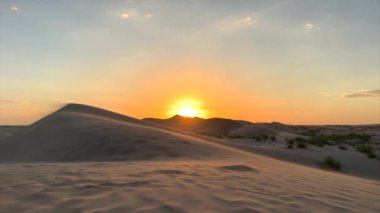 Altın günbatımı çöl kumlarının üzerinde parlıyor, yumuşak turuncu ışık gökyüzüne yayılıyor, koyu kumlarda ılık tonlarda yaratıyor, barışçıl atmosfer doğal güzellikleri, çöl sükunetini vurguluyor,. 