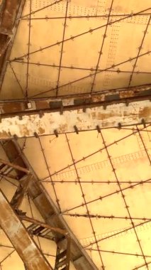 Paslı kirişler, terk edilmiş çöl yapısındaki sütunlar tarafından desteklenen geometrik tavan üzerinde kesişiyor. Endüstriyel bozulma, yıpranmış yüzeyler ve ıssız atmosfer uzun vadede ihmal gösteriyor..
