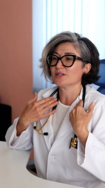 Caucasian doctor with short gray hair, white coat, black glasses gestures passionately during conversation, stethoscope resting around neck, expressing compassion and confidence. Professional empathy. clipart