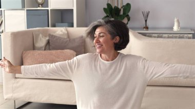 Gray-haired woman, white top, stretches arm with joy, movement suggests playful energy, beige couch in background, peaceful, light-filled space reflects calm, relaxed atmosphere. clipart