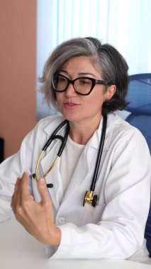 Doctor, Caucasian woman with short gray hair, white coat, gestures confidently while speaking, calm expression, stethoscope resting on shoulders, exuding medical authority. Professional care. clipart